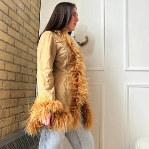 May include: A tan-colored coat with long sleeves and a faux fur trim along the collar, front, and cuffs. The coat is worn over light blue jeans. The coat has a tailored fit and a button closure.
