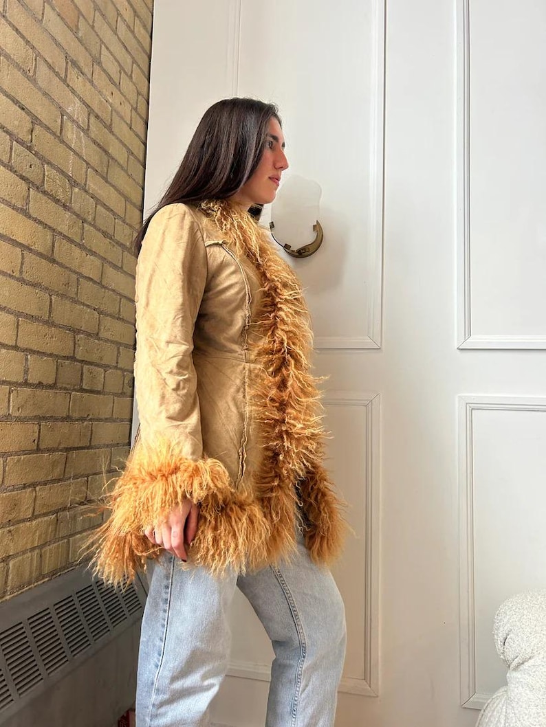 May include: A tan-colored coat with long sleeves and a faux fur trim along the collar, front, and cuffs. The coat is worn over light blue jeans. The coat has a tailored fit and a button closure.