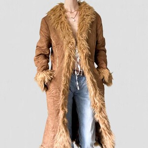 May include: A long, brown coat with faux fur trim on the collar, cuffs, and down the front. The coat has a suede-like texture and a tie closure. The model is wearing blue jeans and a brown belt.