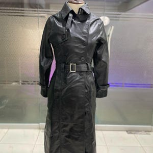 May include: A black leather trench coat on a mannequin. The coat has a double-breasted design, a belt with a silver buckle, and long sleeves. The coat is approximately 140 cm long.