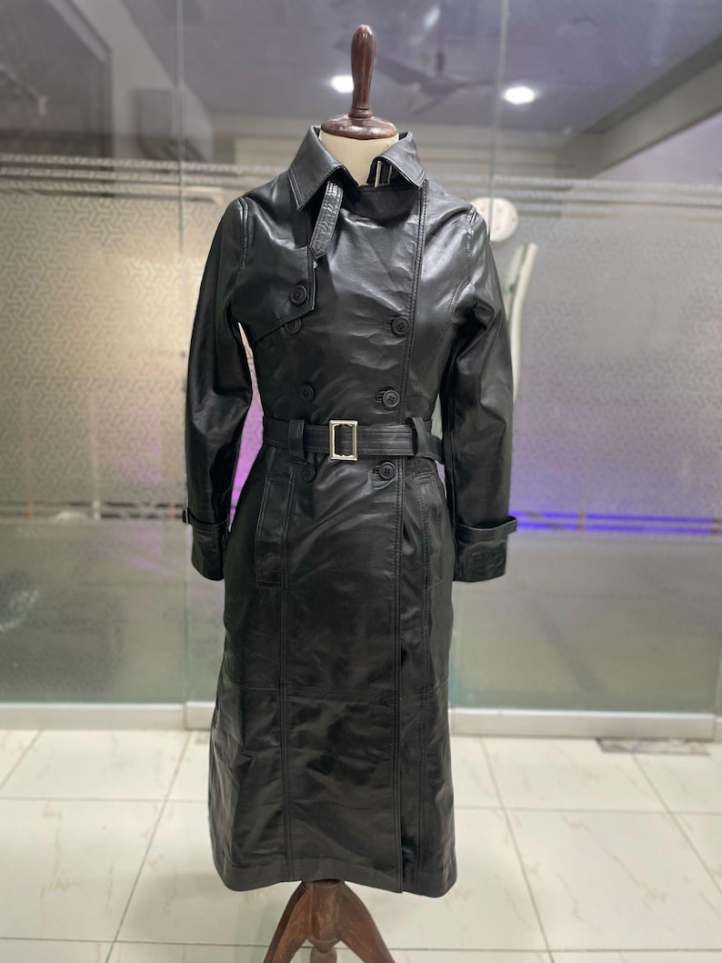 May include: A black leather trench coat on a mannequin. The coat has a double-breasted design, a belt with a silver buckle, and long sleeves. The coat is approximately 140 cm long.