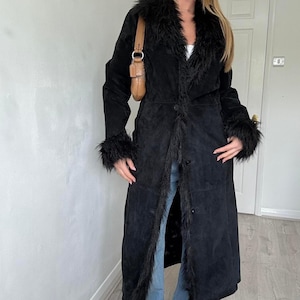 May include: A long black coat with faux fur trim on the collar, cuffs, and down the front. The coat has a button closure and a suede-like texture. A brown shoulder bag is visible.