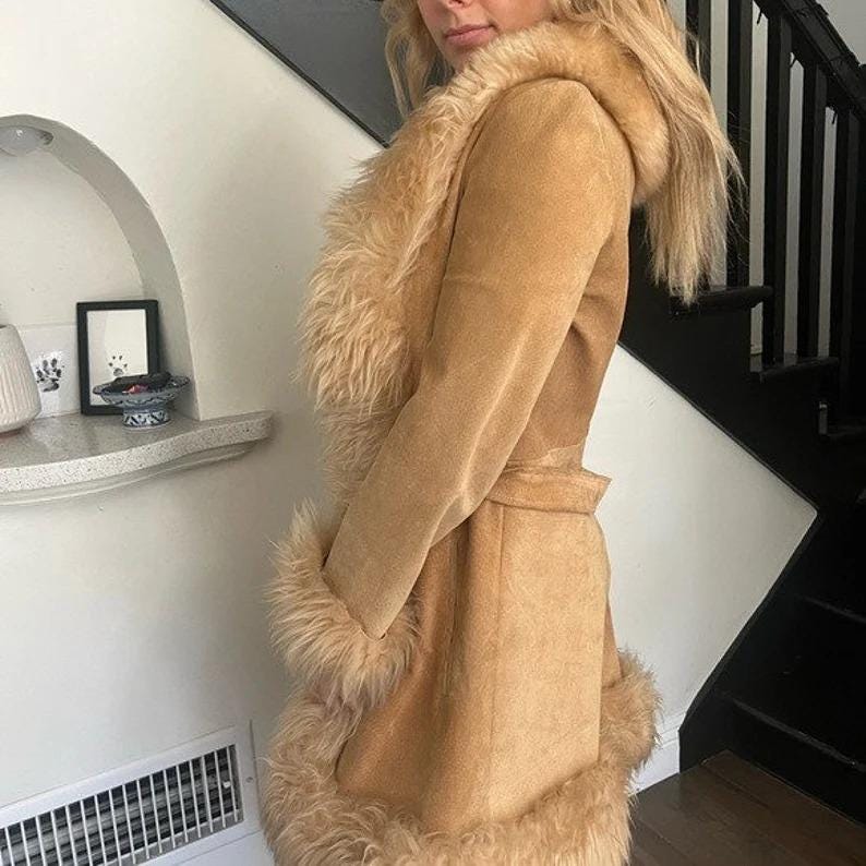 May include: A tan suede coat with a faux fur trim around the collar, cuffs, and hem. The coat has a hood and a belt at the waist. The coat is a light brown color and appears to be made of suede.