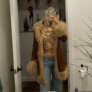 May include: A brown suede coat with tan faux fur trim and cuffs. The coat is open, revealing a patterned crop top and blue jeans. The image is taken in a mirror.