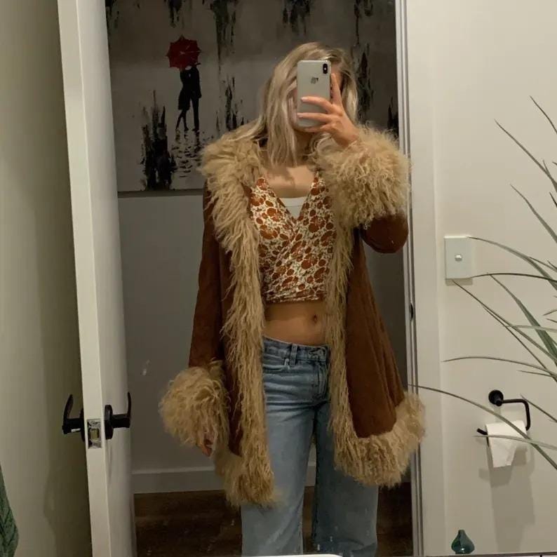 May include: A brown suede coat with tan faux fur trim and cuffs. The coat is open, revealing a patterned crop top and blue jeans. The image is taken in a mirror.