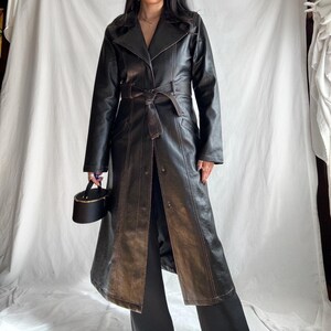 May include: A long black leather trench coat with brown stitching and a belt. The coat has a classic design with a collar, lapels, and a single-breasted closure.