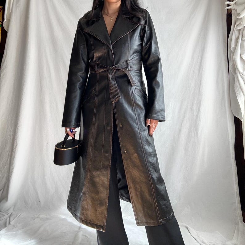 May include: A long black leather trench coat with brown stitching and a belt. The coat has a classic design with a collar, lapels, and a single-breasted closure.