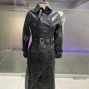 May include: A black leather trench coat on a mannequin. The coat has a double-breasted design, a belt with a silver buckle, and a classic collar. The coat is knee-length and has long sleeves.