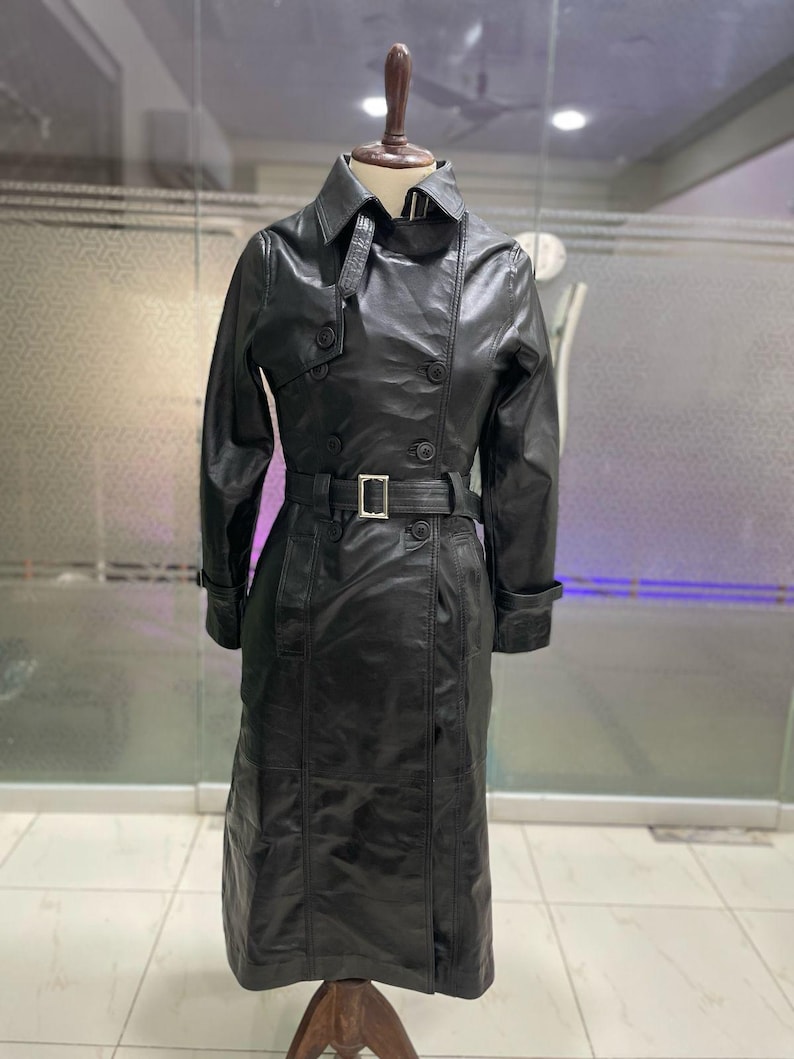 May include: A black leather trench coat on a mannequin. The coat has a double-breasted design, a belt with a silver buckle, and a classic collar. The coat is knee-length and has long sleeves.
