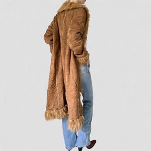 May include: A long brown coat with faux fur trim on the collar, cuffs, and hem. The coat is made of a suede-like material and has a flowing design. The coat is worn with blue jeans and brown boots.