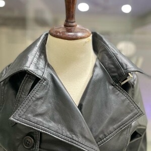 May include: A black leather trench coat with a notched collar and double-breasted button closure. The coat features a belt detail and is displayed on a mannequin. The coat is a classic outerwear piece.