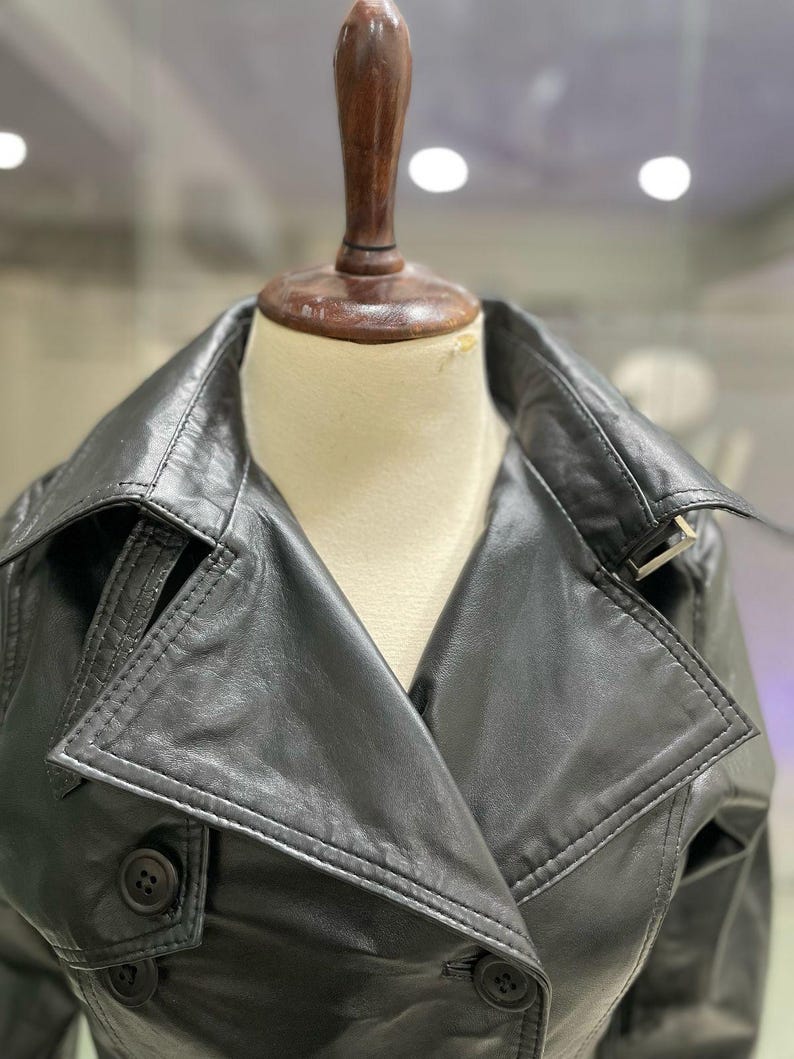 May include: A black leather trench coat with a notched collar and double-breasted button closure. The coat features a belt detail and is displayed on a mannequin. The coat is a classic outerwear piece.