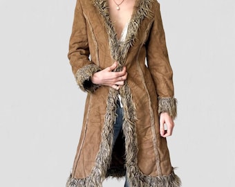 May include: A long brown coat with faux fur trim around the collar, cuffs, and down the front. The coat has a suede-like texture and a tailored fit. The coat is open, revealing a glimpse of a white top and blue jeans.