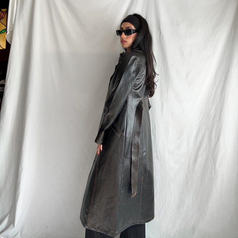 May include: A long black leather trench coat with a belt and a satin lining. The coat has a stand-up collar and long sleeves.