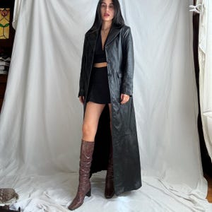 Peut inclure: Un long manteau en cuir noir avec une fermeture boutonnée. Le manteau est porté avec une tenue noire et des bottes hautes marron. Le manteau a un design classique et ajusté.