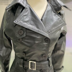 May include: A black leather trench coat with a double-breasted design. The coat has a notched collar, button fastenings, and a belt with a silver buckle. The coat is displayed on a mannequin.