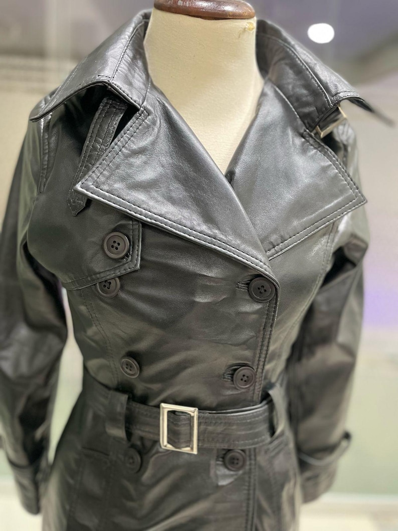 May include: A black leather trench coat with a double-breasted design. The coat has a notched collar, button fastenings, and a belt with a silver buckle. The coat is displayed on a mannequin.