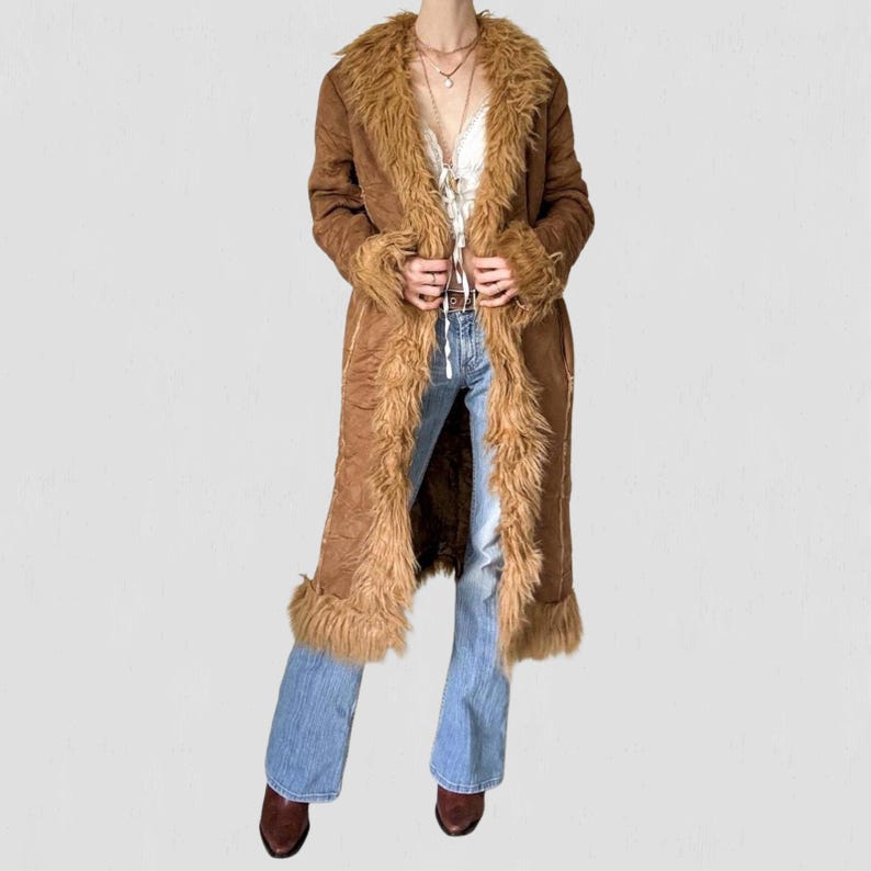May include: A long brown coat with faux fur trim on the collar, cuffs, and edges. The coat is open, revealing a white top and blue jeans. The coat has a vintage, bohemian style.