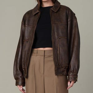 May include: A brown leather bomber jacket with a collared neckline and front pockets. The jacket has a slightly oversized fit and ribbed cuffs and hem. The model is wearing a black top and brown pants.