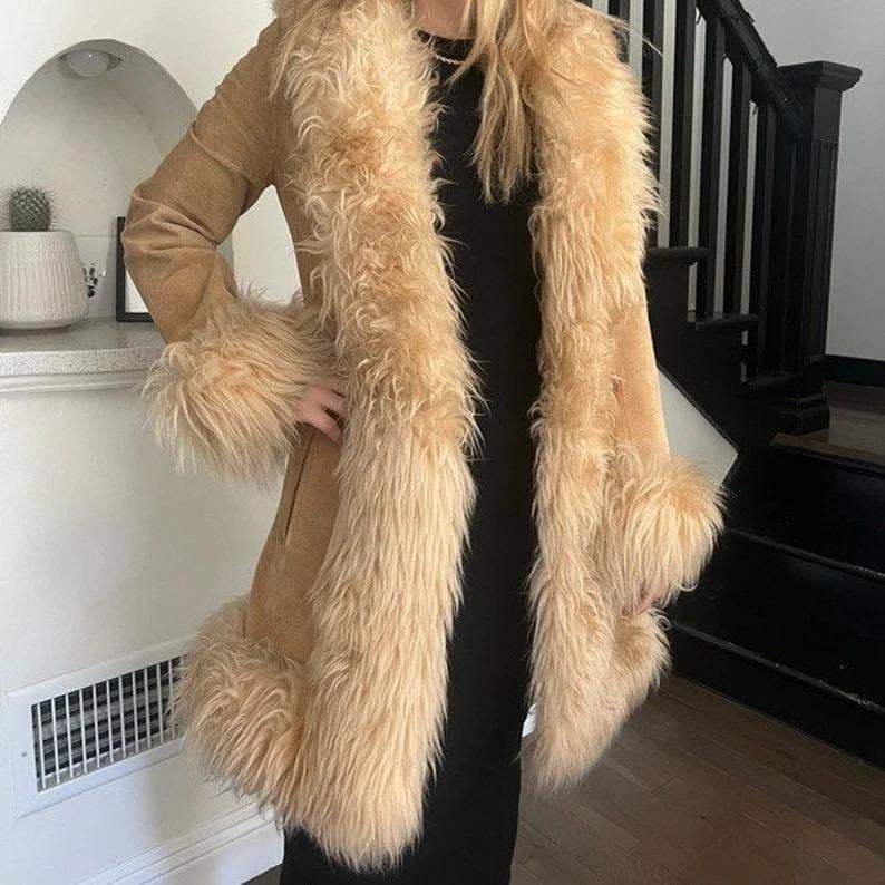 May include: A tan suede coat with a faux fur collar and cuffs. The coat has a long, flowing design with a button closure. The faux fur is a light beige color, and the coat is worn over a black dress.