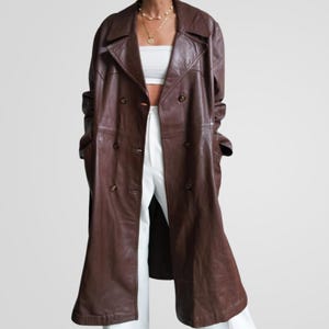 May include: A long, brown leather trench coat with a double-breasted design. The coat features a classic lapel collar, button closures, and pockets. The coat is worn over white pants.
