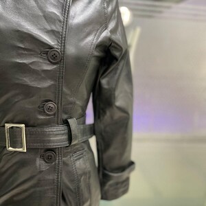 May include: Black leather trench coat with a belted waist and button fastenings. The coat has a classic design with a collar and long sleeves. The belt has a silver buckle.