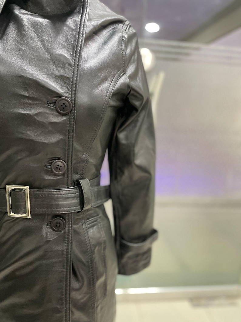 May include: Black leather trench coat with a belted waist and button fastenings. The coat has a classic design with a collar and long sleeves. The belt has a silver buckle.