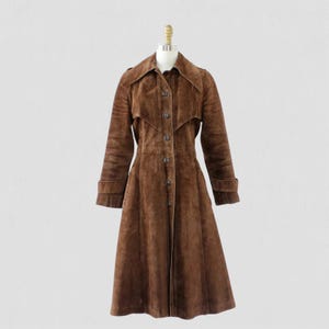 May include: A vintage brown suede trench coat with a button-up front, notched collar, and long sleeves. The coat has a flared skirt and is displayed on a mannequin. A classic outerwear piece.