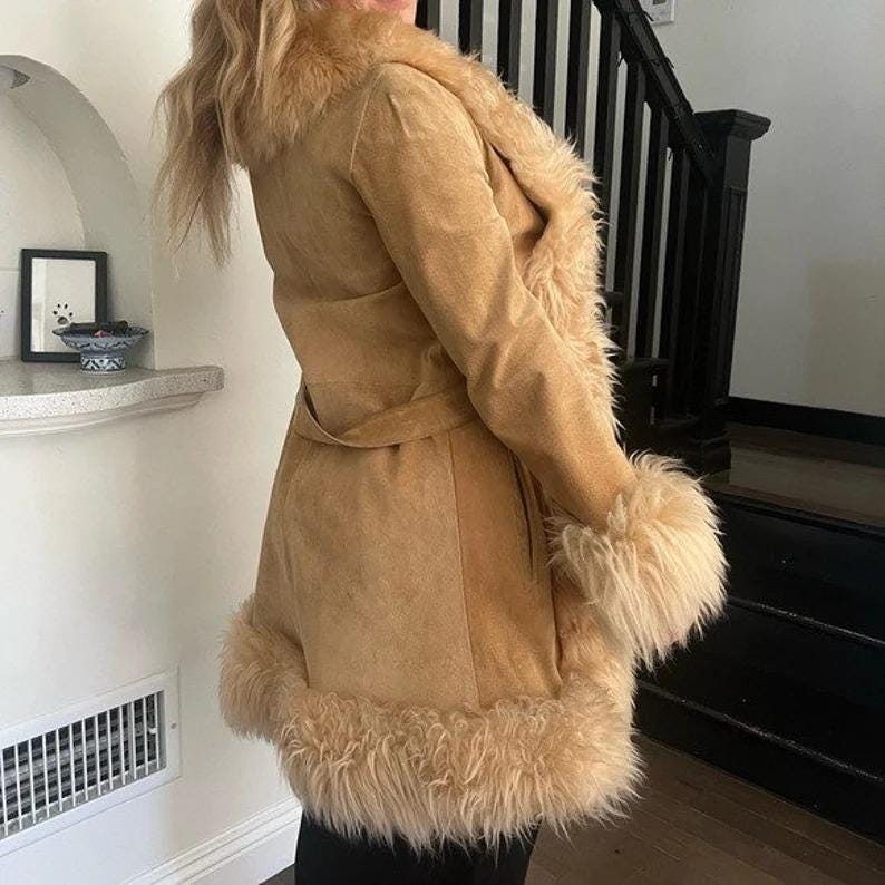 May include: A tan suede coat with a faux fur collar, cuffs, and hem. The coat features a matching belt and a classic silhouette. The fur trim adds a touch of luxury to this vintage-inspired outerwear piece.