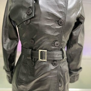 May include: A black leather trench coat with a double-breasted design. The coat has a belt with a silver buckle, multiple buttons, and a classic collar. The leather has a slight sheen.
