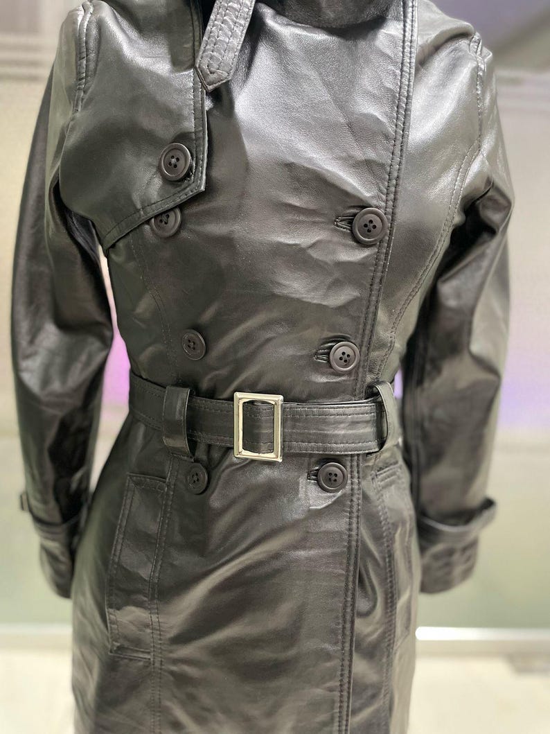 May include: A black leather trench coat with a double-breasted design. The coat has a belt with a silver buckle, multiple buttons, and a classic collar. The leather has a slight sheen.