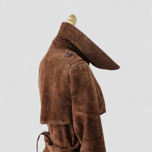 May include: A brown suede trench coat displayed on a mannequin. The coat features a unique, oversized collar, a button detail on the shoulder, and a belted waist. The coat's rich brown color and soft texture are prominent.