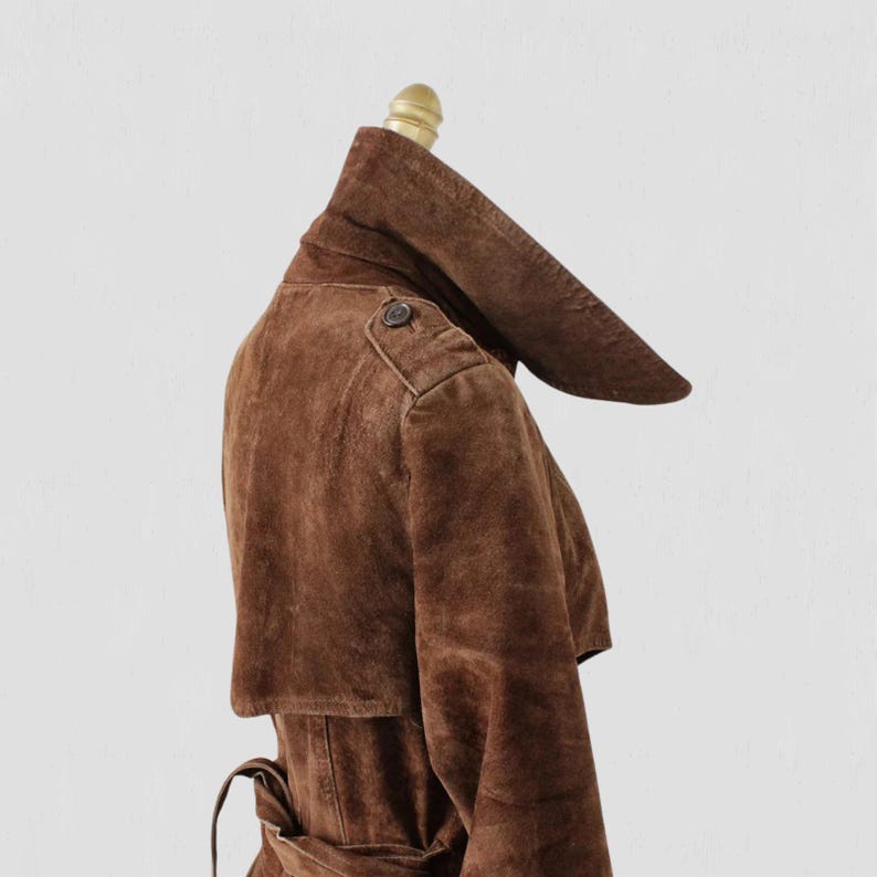 May include: A brown suede trench coat displayed on a mannequin. The coat features a unique, oversized collar, a button detail on the shoulder, and a belted waist. The coat's rich brown color and soft texture are prominent.