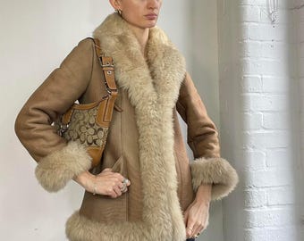 Puede incluir: Un abrigo de borrego color canela con un ribete de piel sintética color crema alrededor del cuello, los puños y el dobladillo. El abrigo tiene un diseño clásico con cierre de botones y un bolso Coach a juego con un estampado marrón y beige.