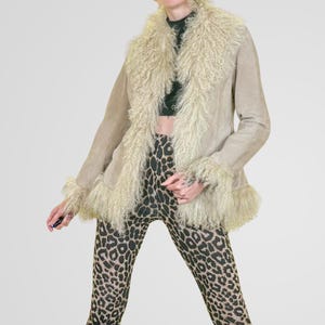 May include: A tan suede jacket with a cream-colored, shaggy fur collar and cuffs. The jacket is open, revealing a black top and leopard print leggings. The jacket has a relaxed fit and a vintage aesthetic.