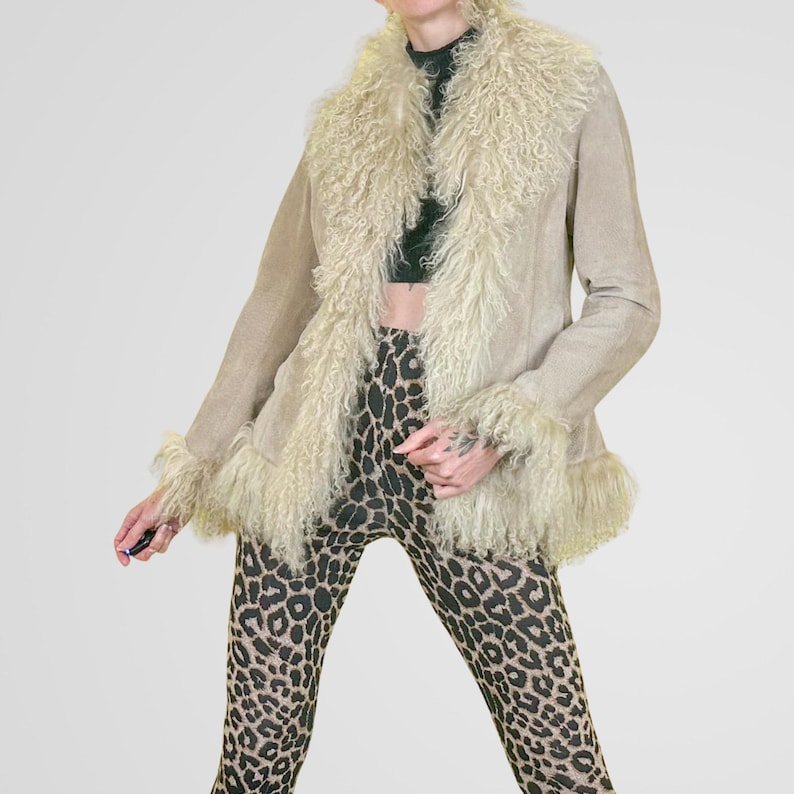 May include: A tan suede jacket with a cream-colored, shaggy fur collar and cuffs. The jacket is open, revealing a black top and leopard print leggings. The jacket has a relaxed fit and a vintage aesthetic.