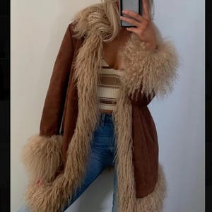 May include: A brown suede coat with a cream-colored faux fur trim on the collar, cuffs, and hem. The coat is open, revealing a striped tank top and blue jeans. The coat has a long, flowing design.