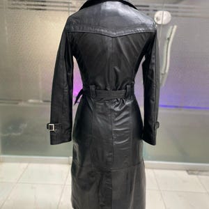 May include: Black leather trench coat with a belted waist and long sleeves. The coat features a classic collar, a button closure, and a knee-length hem. The coat is displayed on a mannequin. The coat has a sleek, polished appearance.