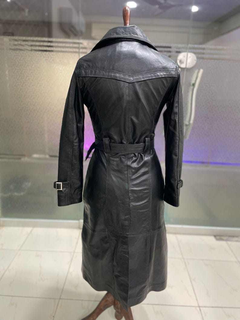 May include: Black leather trench coat with a belted waist and long sleeves. The coat features a classic collar, a button closure, and a knee-length hem. The coat is displayed on a mannequin. The coat has a sleek, polished appearance.