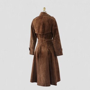 May include: A brown suede trench coat displayed on a mannequin. The coat features a belted waist, long sleeves, and a flared skirt. The coat has a classic trench coat design with a collar and button details. The coat is a medium brown color.