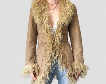 May include: A tan suede coat with a shaggy, light brown fur collar and trim. The coat has long sleeves, a tailored fit, and two front pockets. The model is wearing blue jeans.