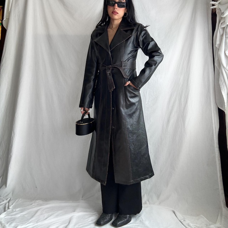May include: A long black leather trench coat with brown stitching and a belt. The coat has a classic design with a collar, lapels, and pockets.