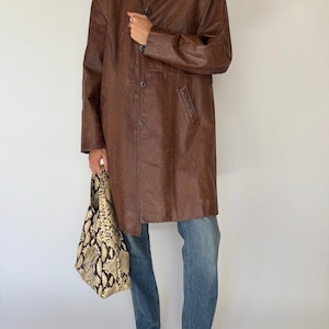 May include: A brown leather coat with a button-up front, worn with blue jeans and black shoes. The coat is knee-length and has a classic collar. A snakeskin-patterned tote bag is held in the hand.
