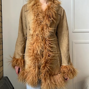 May include: A tan suede coat with a shaggy, light brown fur trim around the collar, cuffs, and down the front. The coat has long sleeves and a tailored fit. The fur trim adds a vintage, bohemian flair to the outerwear.