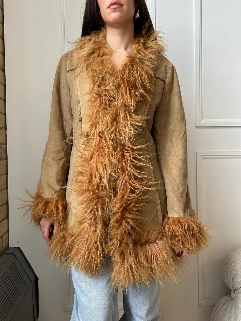 May include: A tan suede coat with a shaggy, light brown fur trim around the collar, cuffs, and down the front. The coat has long sleeves and a tailored fit. The fur trim adds a vintage, bohemian flair to the outerwear.