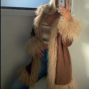 May include: A brown faux fur coat with long sleeves and a shaggy trim. The coat has a long, tailored silhouette and is paired with a striped top and blue jeans. The coat's trim is a light beige color.