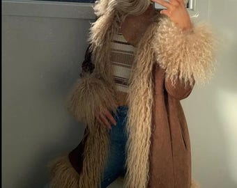 May include: A brown faux fur coat with long sleeves and a shaggy trim. The coat has a long, tailored silhouette and is paired with a striped top and blue jeans. The coat's trim is a light beige color.
