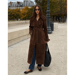 May include: A long, dark brown suede trench coat with a matching waist tie, worn over dark blue denim jeans. The outfit is paired with dark brown ballet flats and a large black leather shoulder bag. The coat features a wide collar and lapels.