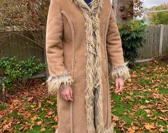 May include: A long, tan coat with faux fur trim on the collar, cuffs, and hem. The coat has a suede-like texture and a tailored fit. The fur trim is a mix of light and dark brown. The coat is worn outdoors on a grassy lawn.