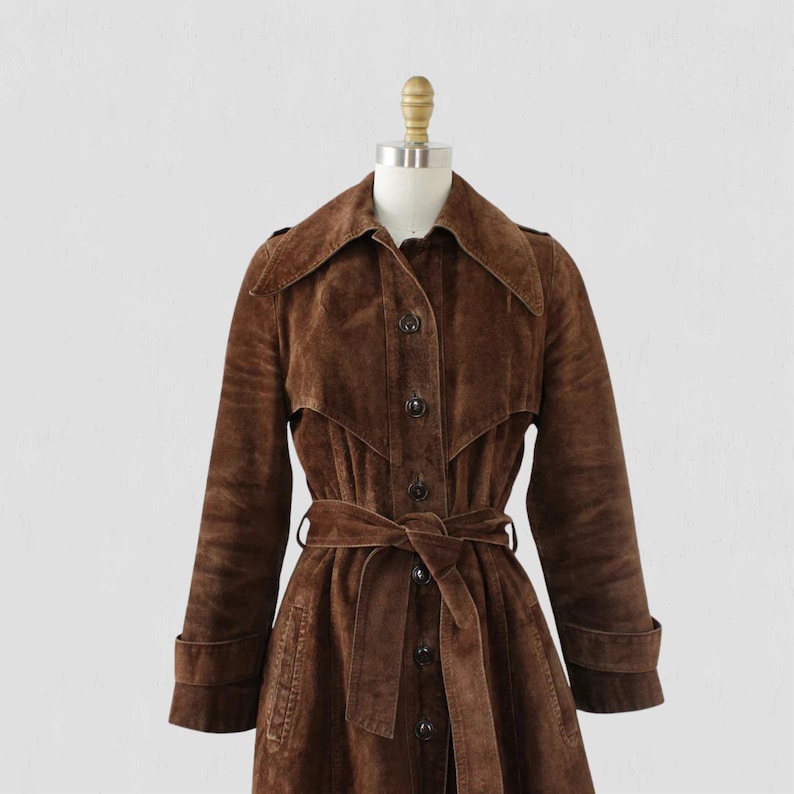 May include: A brown suede trench coat with a belted waist and button-up front. The coat features a wide collar, long sleeves, and is displayed on a mannequin. The coat's length is mid-calf.
